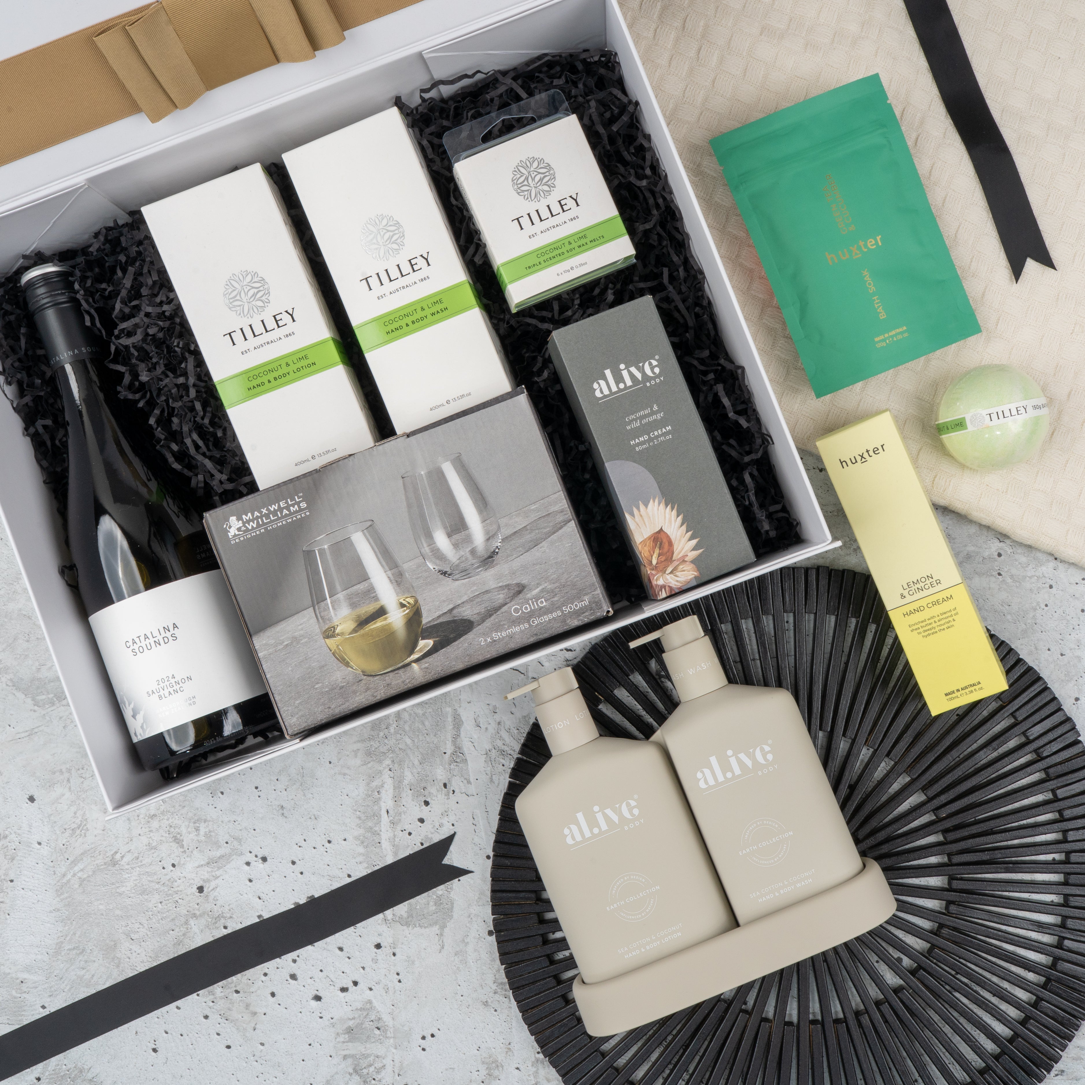 The Hamper Boutique Co's Elegance and Self-Care Gift Hamper includes wine, Tilley soaps, a candle, stemless wine glasses, hand and body lotion, skincare packets—all beautifully arranged on a decorative black mat and cream cloth.