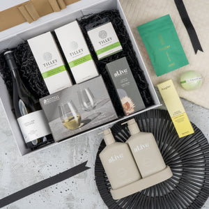 The Hamper Boutique Co's Elegance and Self-Care Gift Hamper includes wine, Tilley soaps, a candle, stemless wine glasses, hand and body lotion, skincare packets—all beautifully arranged on a decorative black mat and cream cloth.