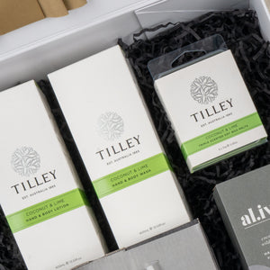 A close-up of The Hamper Boutique Co's Elegance and Self-Care Gift Hamper, featuring Tilley coconut & lime hand & body lotion, hand & body wash, and soy wax melts, beautifully arranged on black shredded paper.
