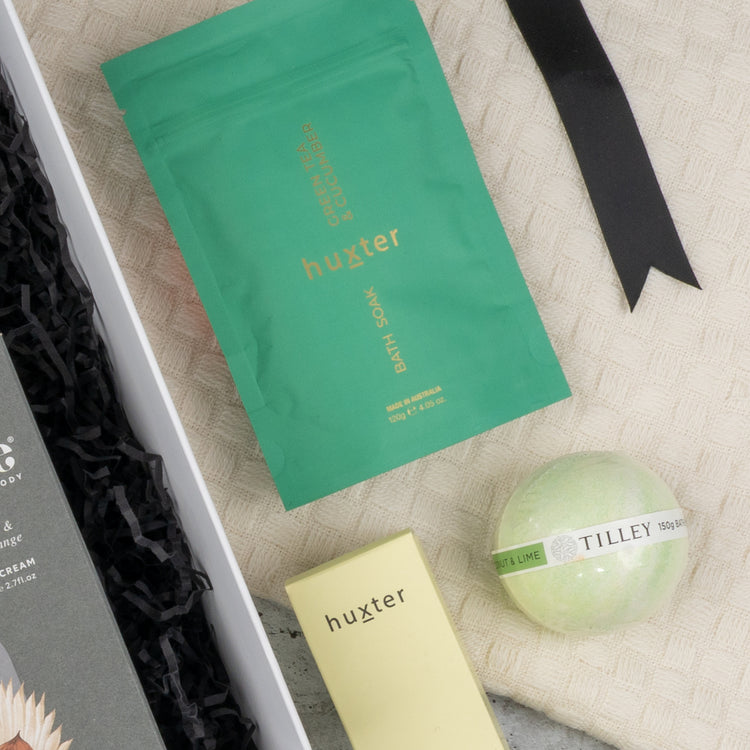 A flat lay of The Hamper Boutique Co’s Elegance and Self-Care Gift Hamper featuring a green bath soak, yellow box, round green bath bomb, and a beige textured towel tied with a black ribbon.
