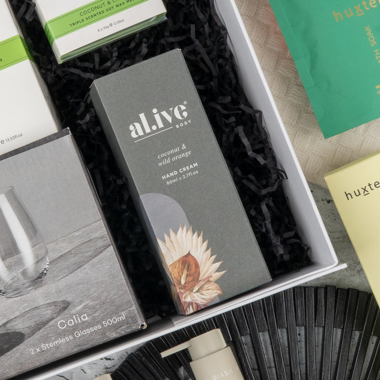 The Hamper Boutique Co's Elegance and Self-Care Gift Hamper includes Alive Body coconut & wild orange hand cream, a Calla set of 2 stemless glasses, and assorted gifts beautifully arranged on black shredded paper.