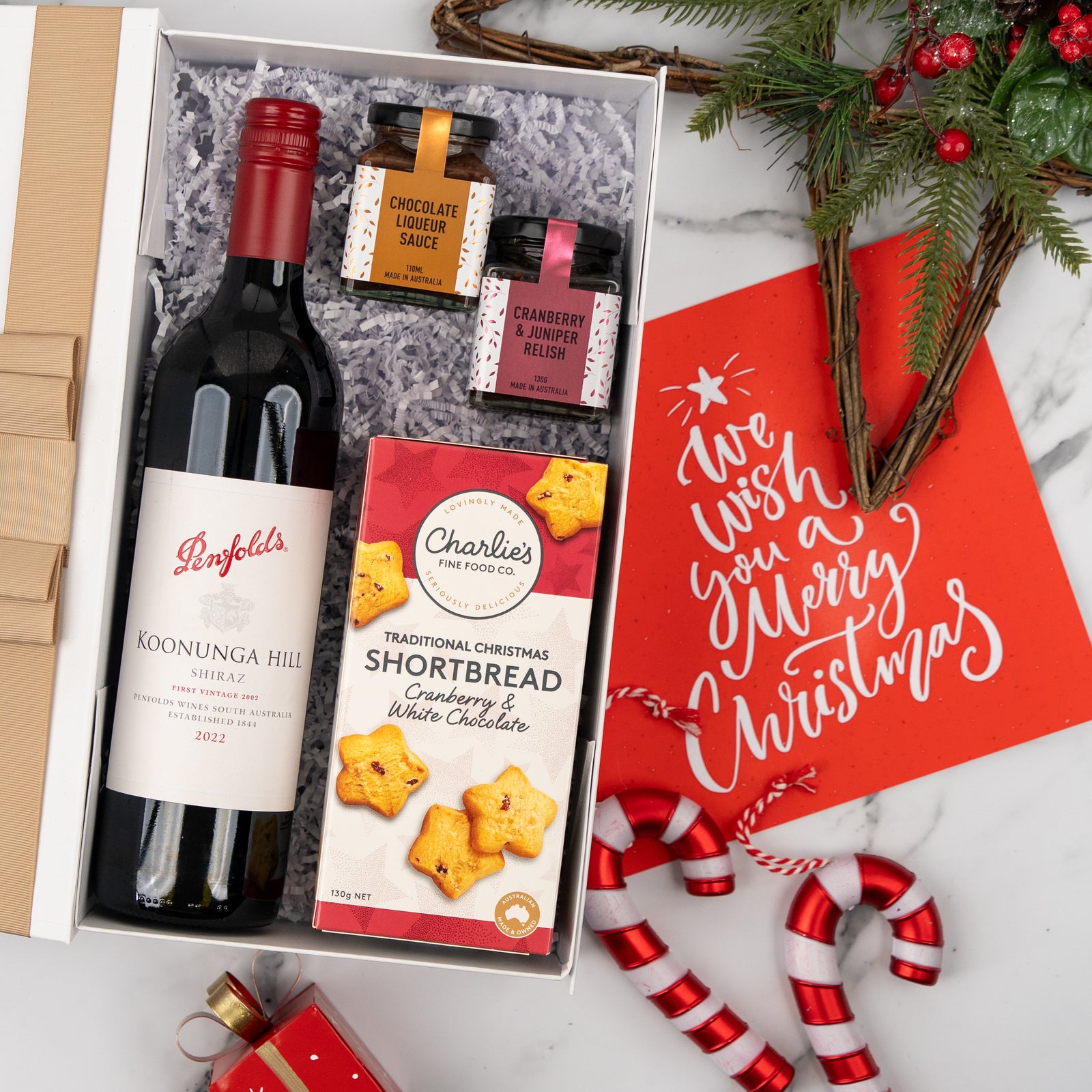The Ruby and Relish Rejoice by The Hamper Boutique Co is a festive gift box featuring wine, chocolate liqueur sauce, relish, and shortbread. Beside it sits a red "We wish you a Merry Christmas" card, candy canes, ornaments, and holiday greenery.