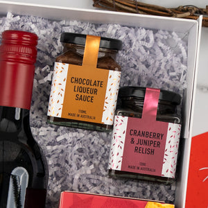 Ruby & Relish Rejoice by The Hamper Boutique Co is a gift box with shredded paper, featuring a bottle of red wine, a jar of chocolate liqueur sauce, and a jar of cranberry & juniper relish with colorful labels, all neatly arranged.