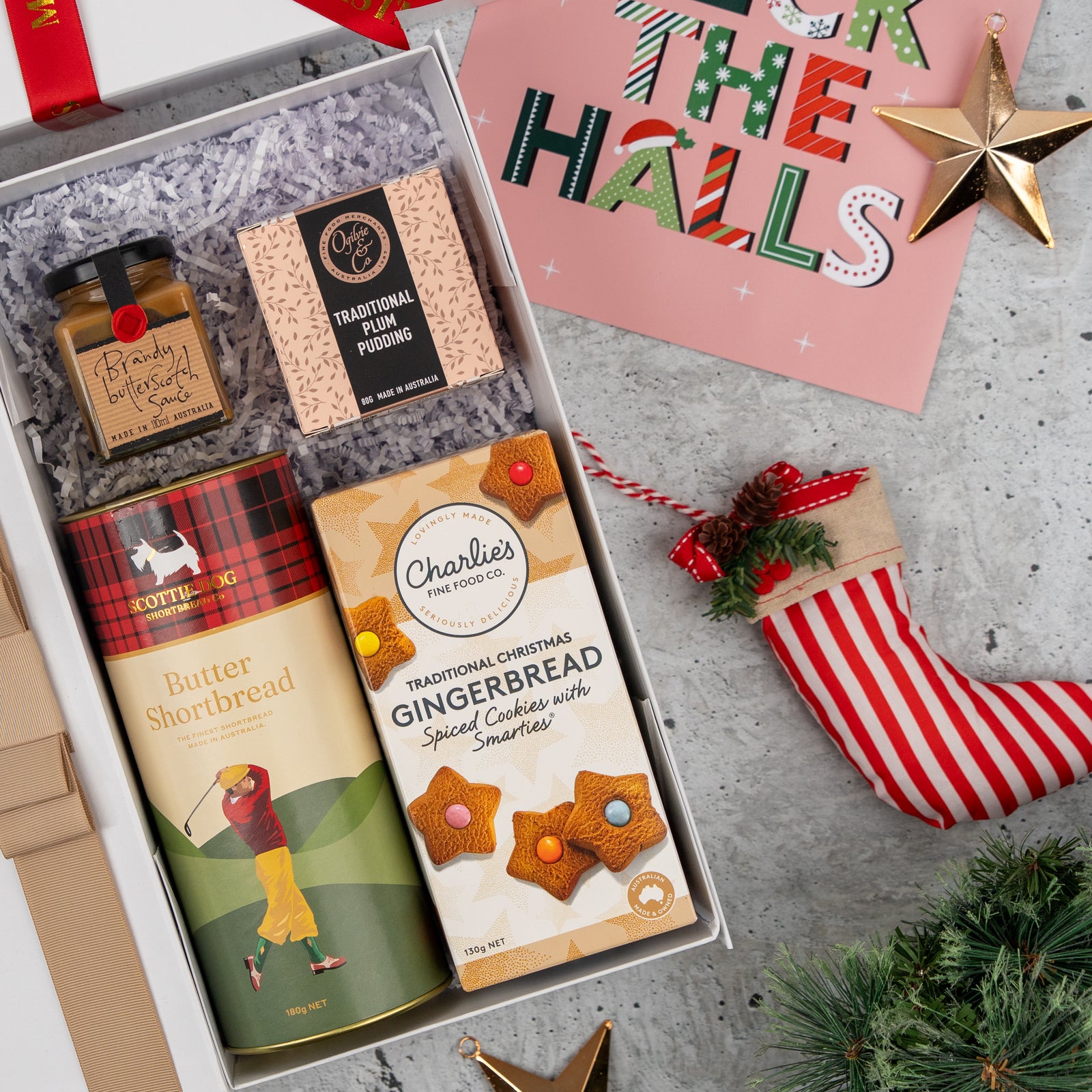 The Sweet Traditions gift box by The Hamper Boutique Co, featuring festive treats, sits beside a striped stocking, gold star ornaments, and a pink "Deck the Halls" card on a gray surface.