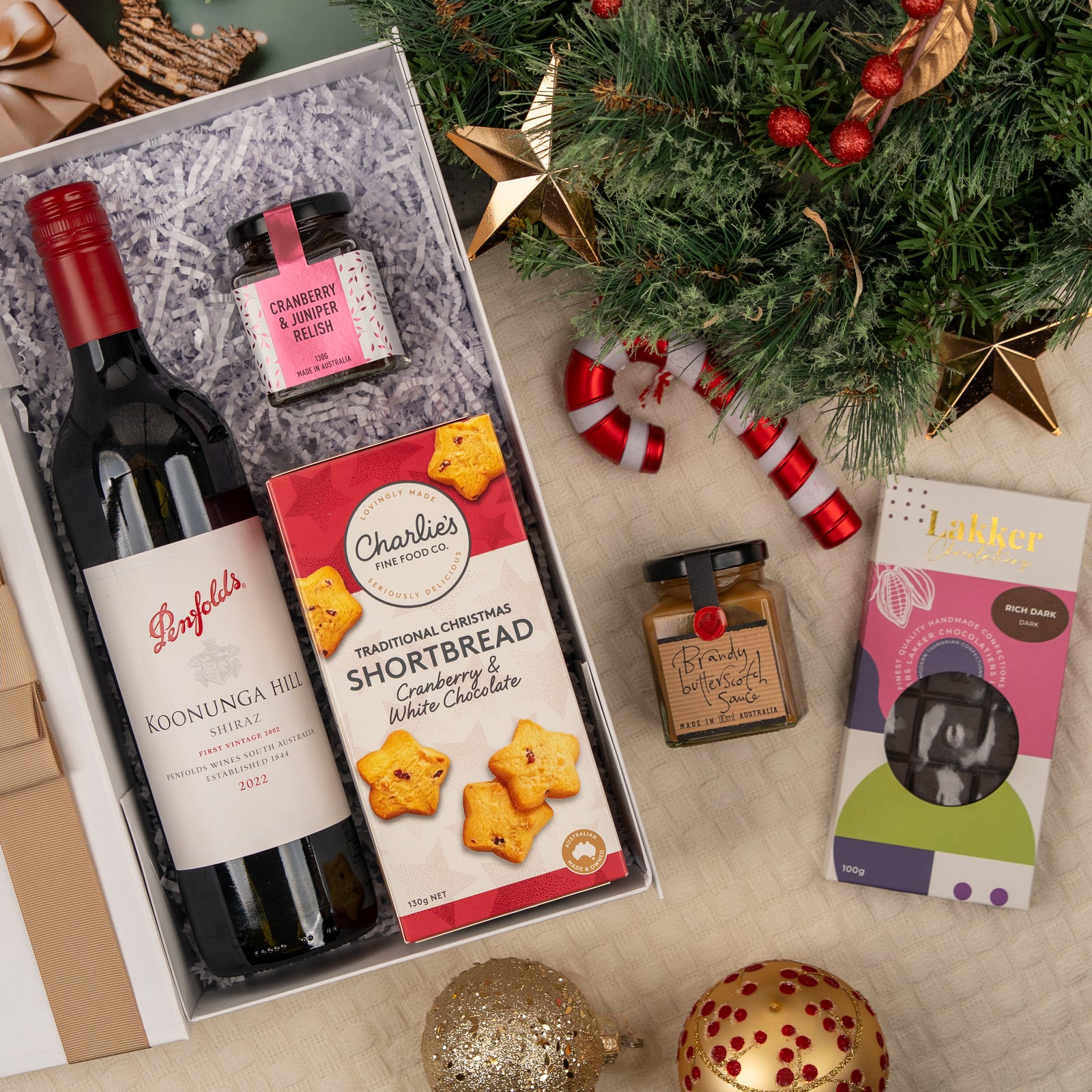 Merry Shiraz Moments by The Hamper Boutique Co features Penfolds red wine, cranberry relish, Christmas shortbread, chocolate fudge sauce, and a chocolate bar—perfectly presented beside a decorated Christmas tree and ornaments.