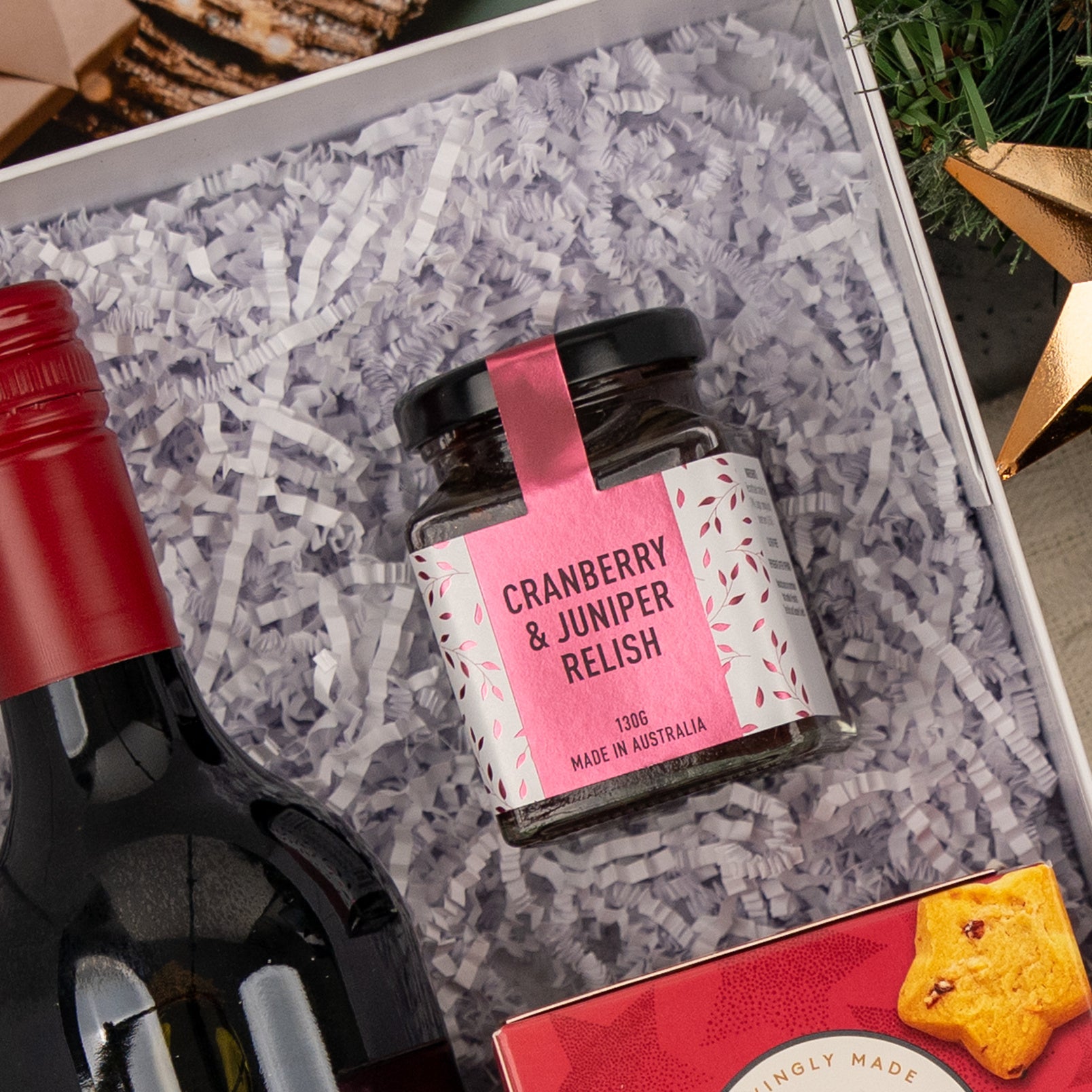 Merry Shiraz Moments by The Hamper Boutique Co features a jar of Cranberry & Juniper Relish, a bottle of red wine, and assorted snacks, all elegantly presented in white shredded paper.