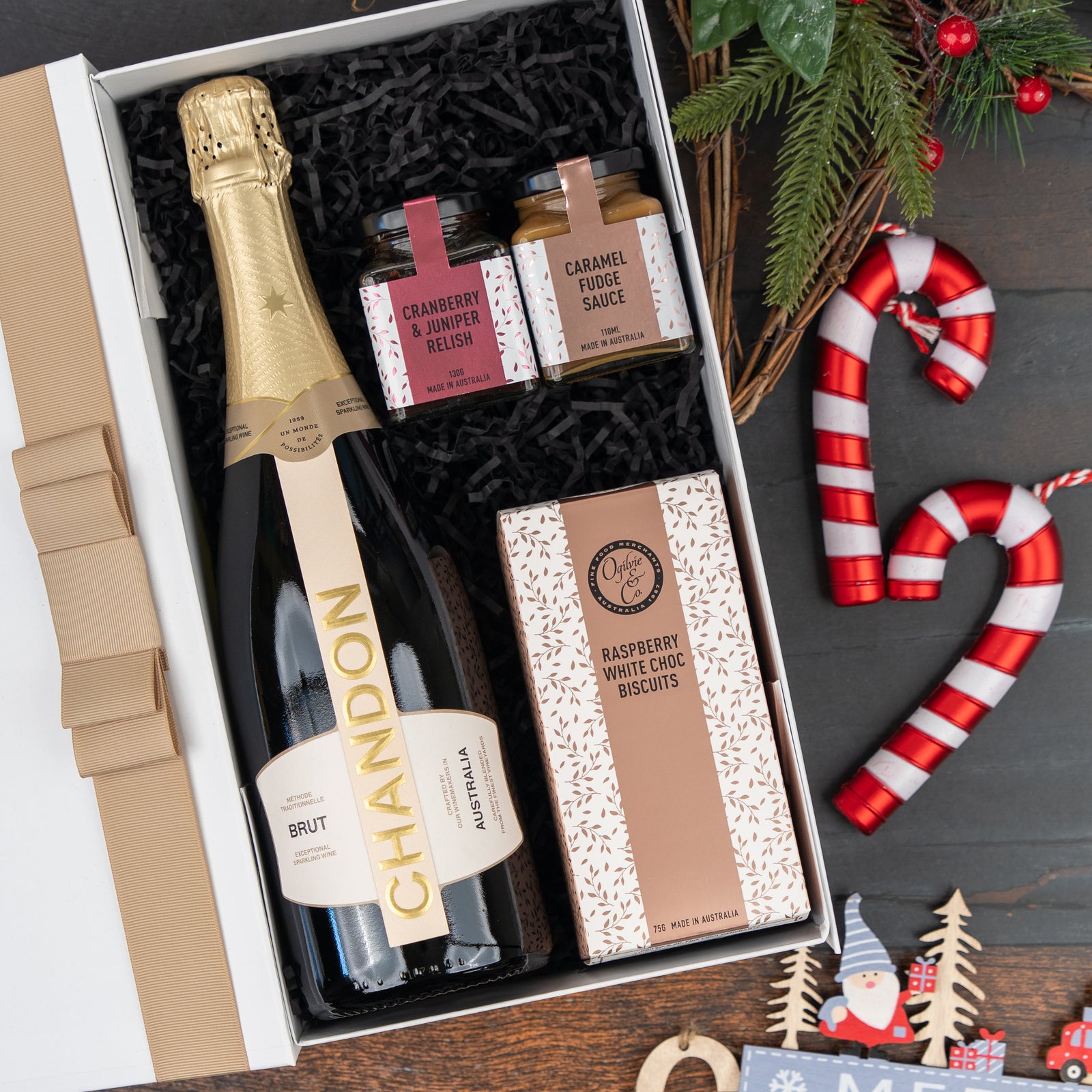 The Hamper Boutique Co’s Chandon Sparkling Gourmet Hamper features a bottle of Chandon, cranberry & juniper relish, caramel fudge sauce, raspberry white choc biscuits, and festive decor like candy canes, pine branches, and ornaments.