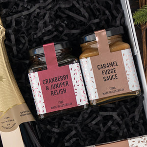 The Hamper Boutique Co’s Chandon Sparkling Gourmet Hamper features jars of “Cranberry & Juniper Relish” and “Caramel Fudge Sauce” with decorative labels, nestled in black crinkle paper alongside a bottle of champagne.