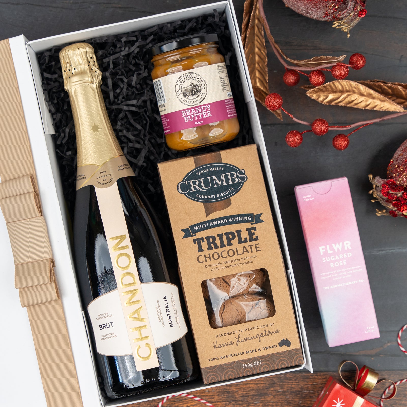 The Hamper Boutique Co’s “Festive Fizz and Pamper” gift box features Chandon Brut, brandy butter, triple chocolate biscuits, and a pink FLWR box, beautifully arranged with festive ornaments on a dark surface.