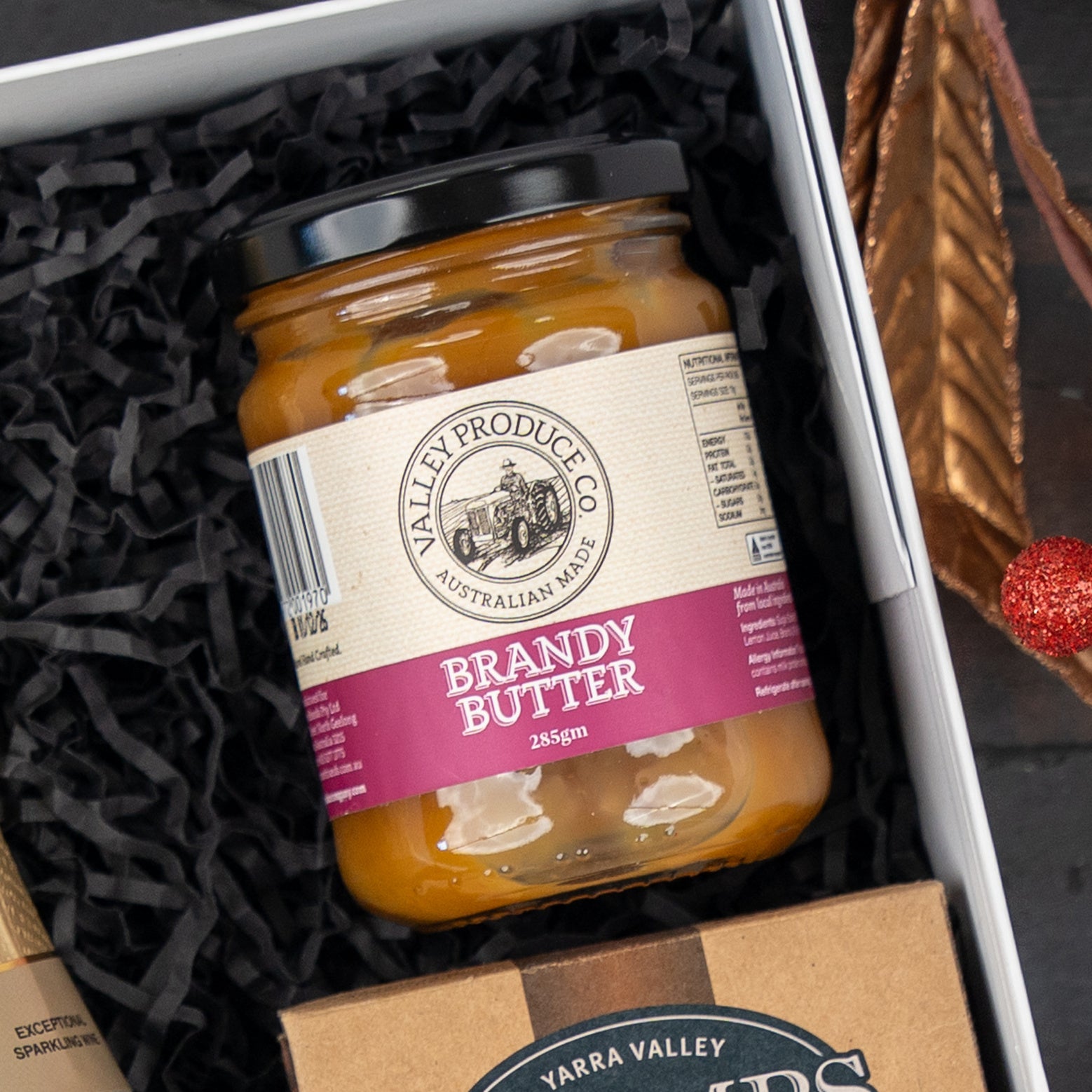 A jar from The Hamper Boutique Co’s Festive Fizz & Pamper set with a black lid sits in a gift box filled with black shredded paper. The label is beige and magenta, and part of another gift item appears beside the jar.