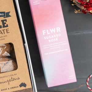 A close-up of The Hamper Boutique Co’s Festive Fizz & Pamper gift set, featuring a pink FLWR Sugared Rose hand cream box beside a brown kraft chocolate treats box on a dark surface with red and white festive décor in the corner.