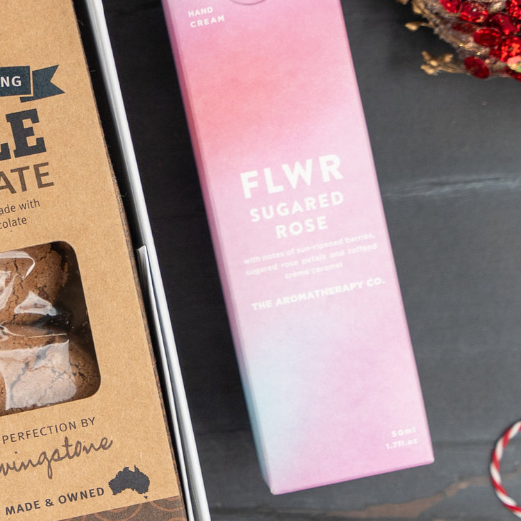 A close-up of The Hamper Boutique Co’s Festive Fizz & Pamper gift set, featuring a pink FLWR Sugared Rose hand cream box beside a brown kraft chocolate treats box on a dark surface with red and white festive décor in the corner.