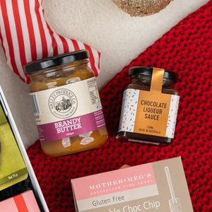A Gourmet Gathering gift set by The Hamper Boutique Co is displayed on a red knitted surface beside a striped item and a Mother Megs gluten-free choco chip biscuit box.