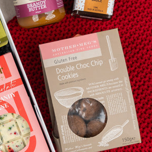The Hamper Boutique Co's Gourmet Gathering is displayed on red knitted fabric, featuring gluten-free double choc chip cookies, jars of brandy butter and marmalade, plus a white chocolate bar with festive red and green toppings.