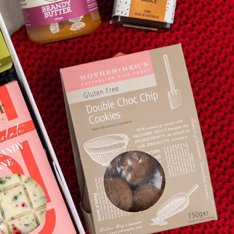 The Hamper Boutique Co's Gourmet Gathering is displayed on red knitted fabric, featuring gluten-free double choc chip cookies, jars of brandy butter and marmalade, plus a white chocolate bar with festive red and green toppings.