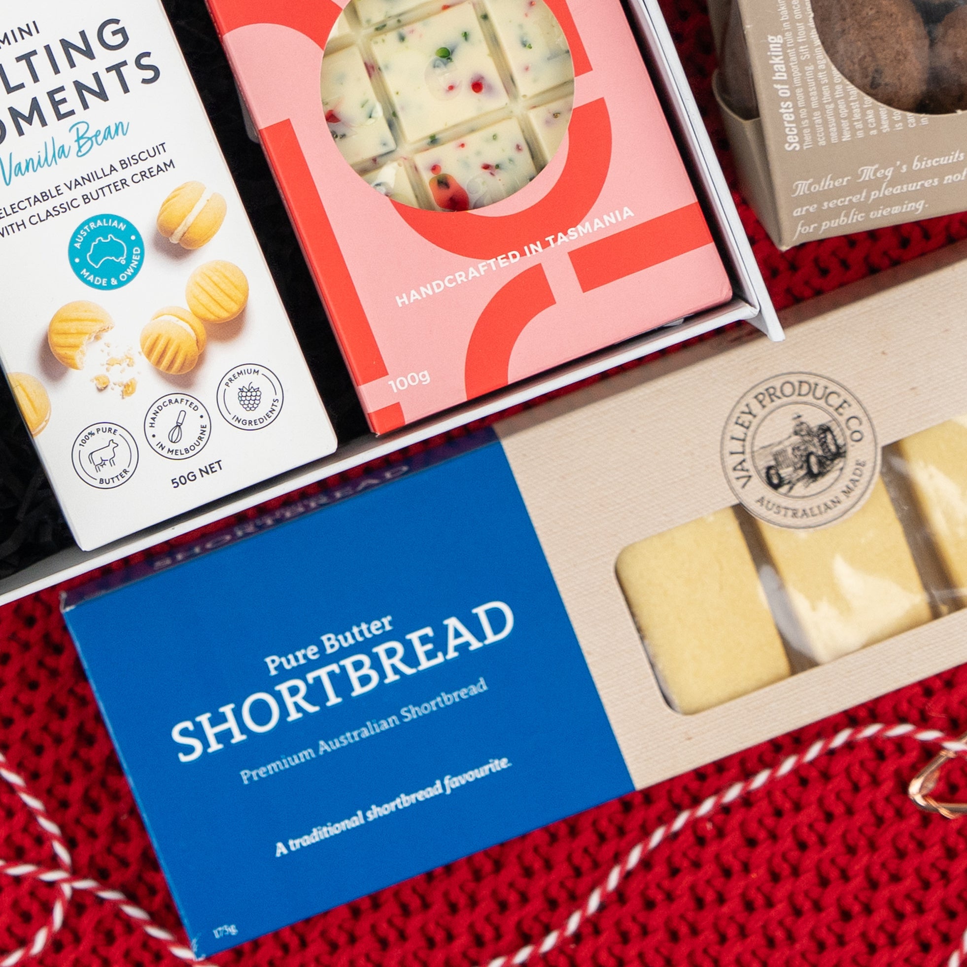 The Hamper Boutique Co's Gourmet Gathering features vanilla cream biscuits, white chocolate with fruit pieces, and pure butter shortbread, artfully arranged on a red knit surface.