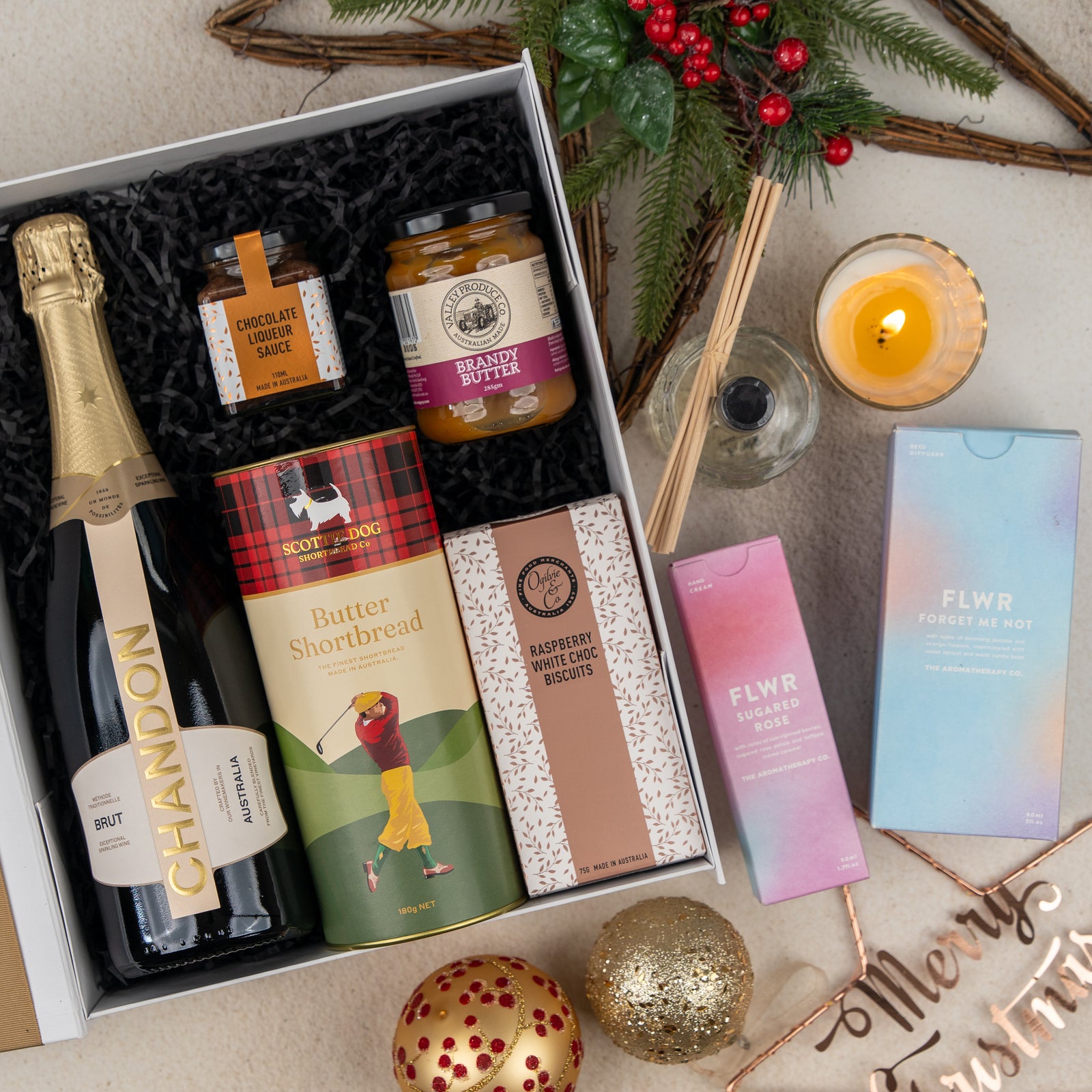 Introducing the Sparkling Comfort and Joy gift box from The Hamper Boutique Co, featuring Chandon Brut, chocolate sauce, brandy butter, shortbread, biscuits, a candle, and two FLWR products. Surrounded by Christmas ornaments, festive décor, and a Merry Christmas sign for the perfect holiday presentation.