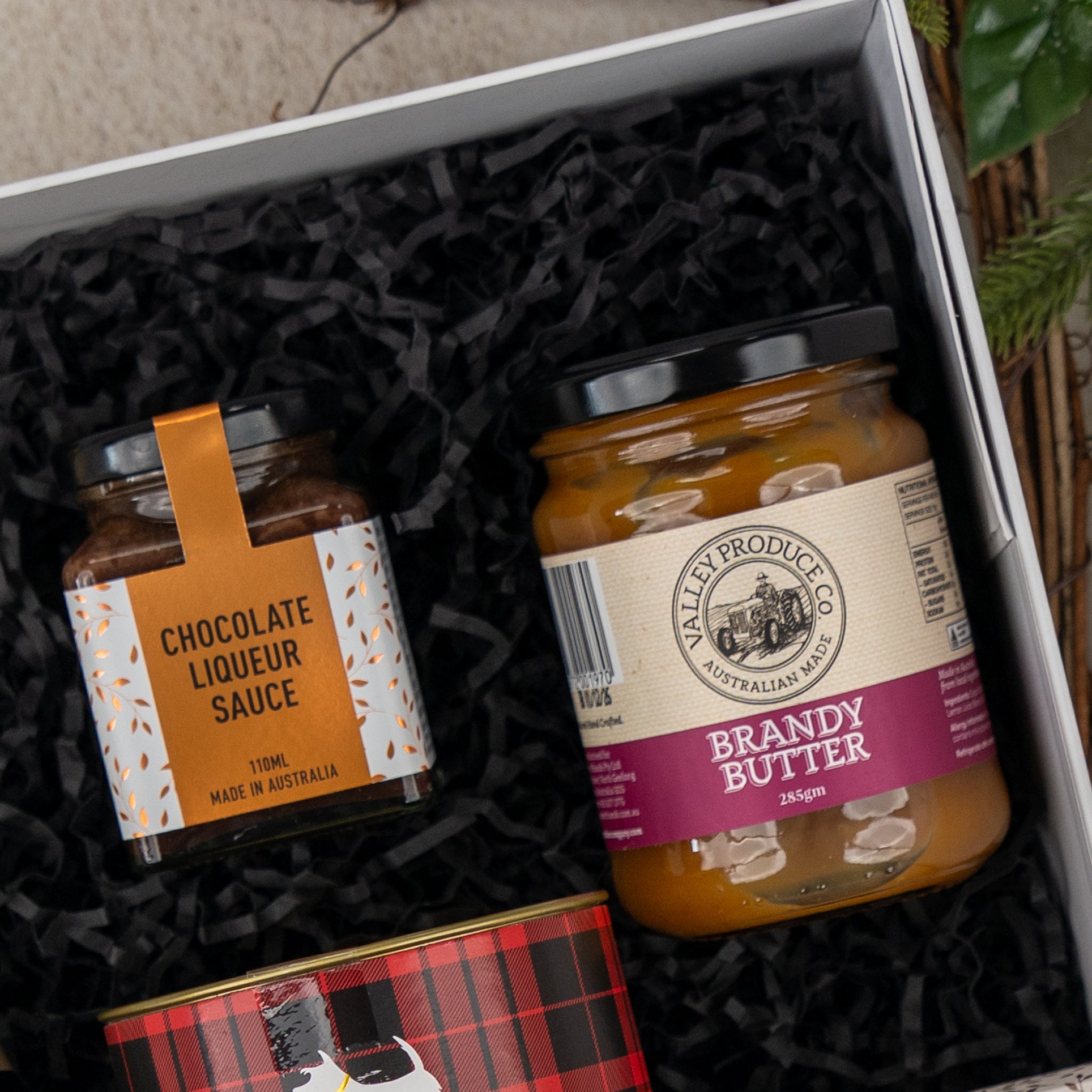 The Hamper Boutique Co’s Sparkling Comfort & Joy gift box features black shredded paper, a jar of chocolate liqueur sauce, a jar of brandy butter, and a glimpse of a red plaid-patterned tin at the bottom.