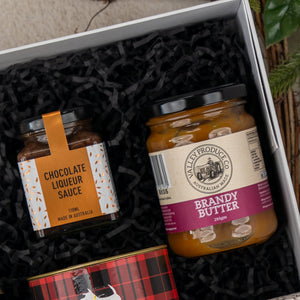 The Hamper Boutique Co’s Sparkling Comfort & Joy gift box features black shredded paper, a jar of chocolate liqueur sauce, a jar of brandy butter, and a glimpse of a red plaid-patterned tin at the bottom.