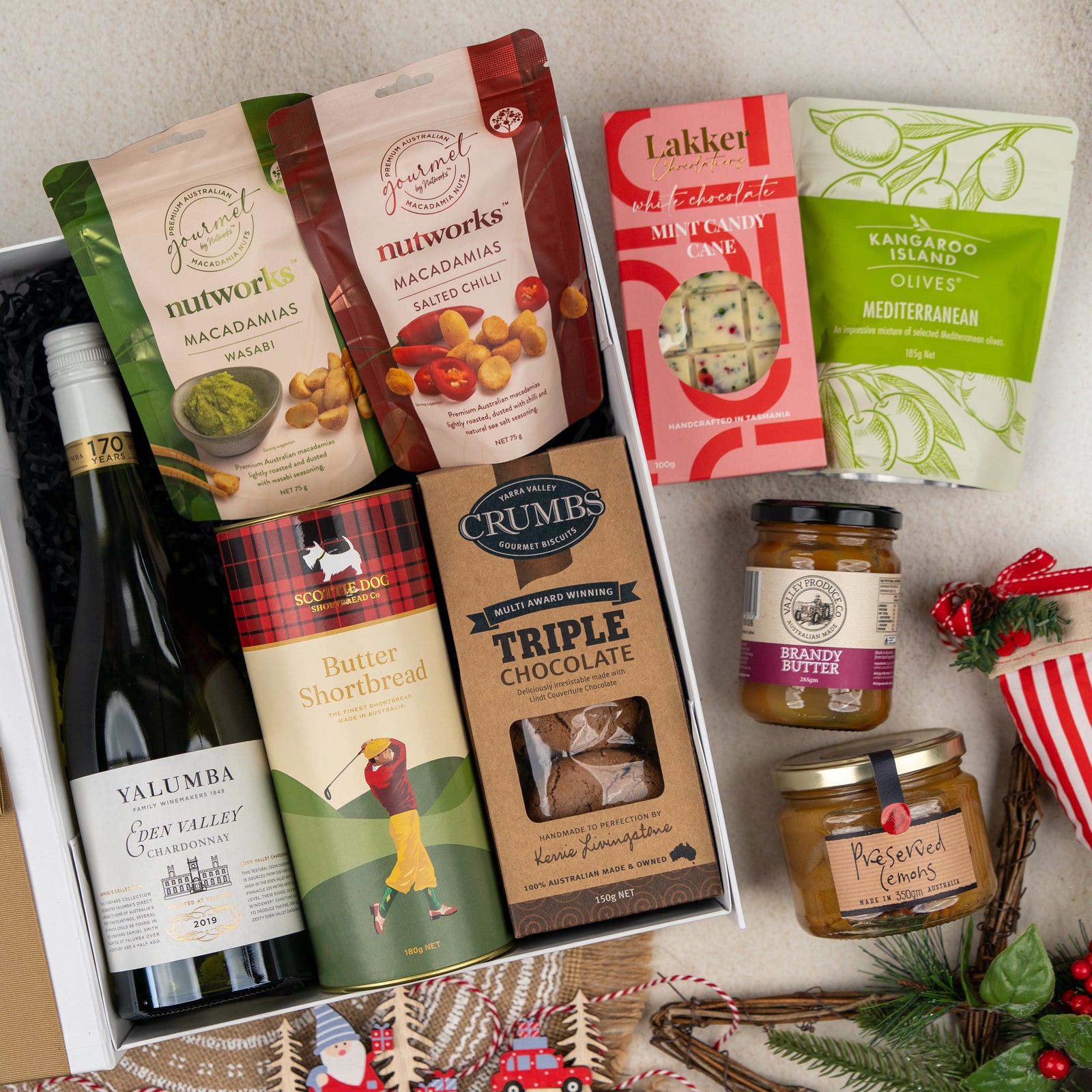 The Gourmet Feast and Chardonnay gift box by The Hamper Boutique Co includes wine, assorted nuts, olives, butter shortbread, triple chocolate biscuits, mint candy, brandy butter, and preserves. Festive decorations surround the box on a neutral background.