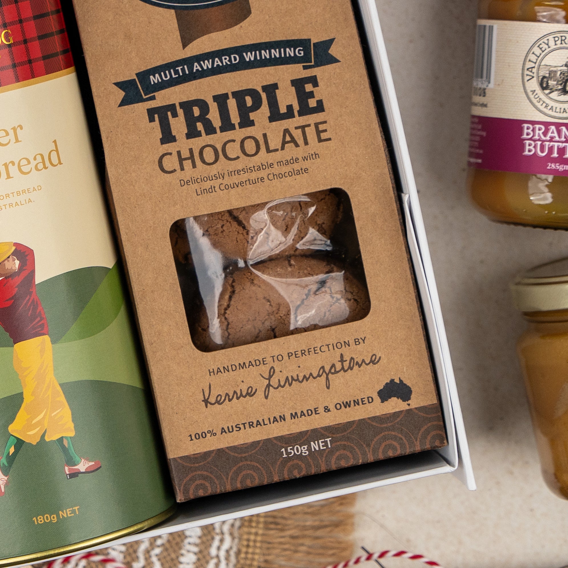 A close-up of The Hamper Boutique Co's Gourmet Feast & Chardonnay gift box containing gingerbread, triple chocolate cookies with a window, honey, and peanut butter on a light-colored surface.