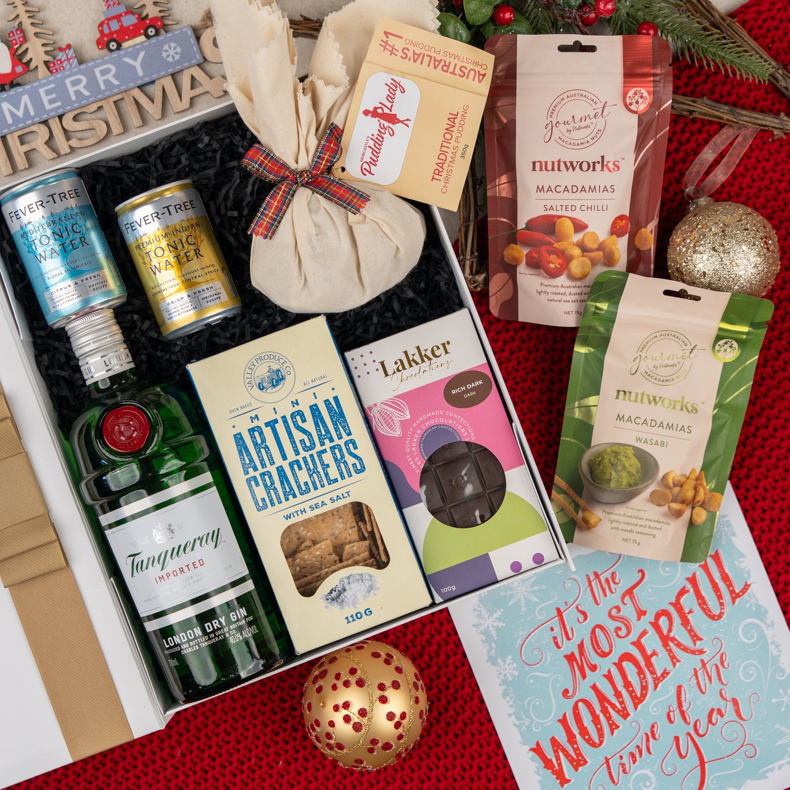 The Cheers and Tonic gift box by The Hamper Boutique Co features Tanqueray gin, Fever-Tree tonic water, crackers, chocolates, macadamias, a Christmas ornament, a holiday card, and festive decorations presented on a vibrant red surface.