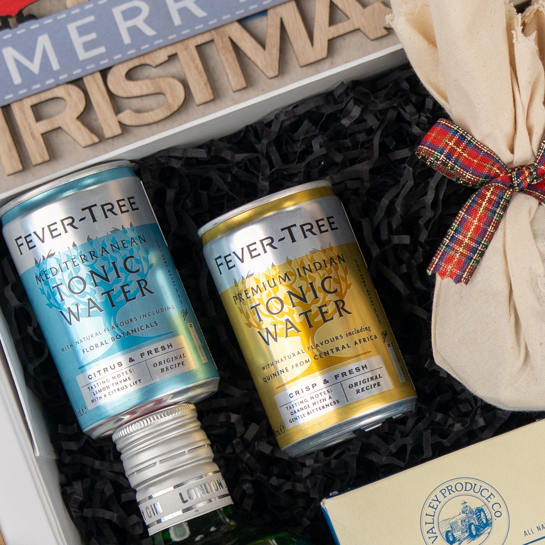 The Hamper Boutique Co's Cheers & Tonic gift box features two Fever-Tree tonic cans—one blue, one gold—in black shredded paper with a plaid ribbon and a wooden Merry Christmas sign peeking out at the top.