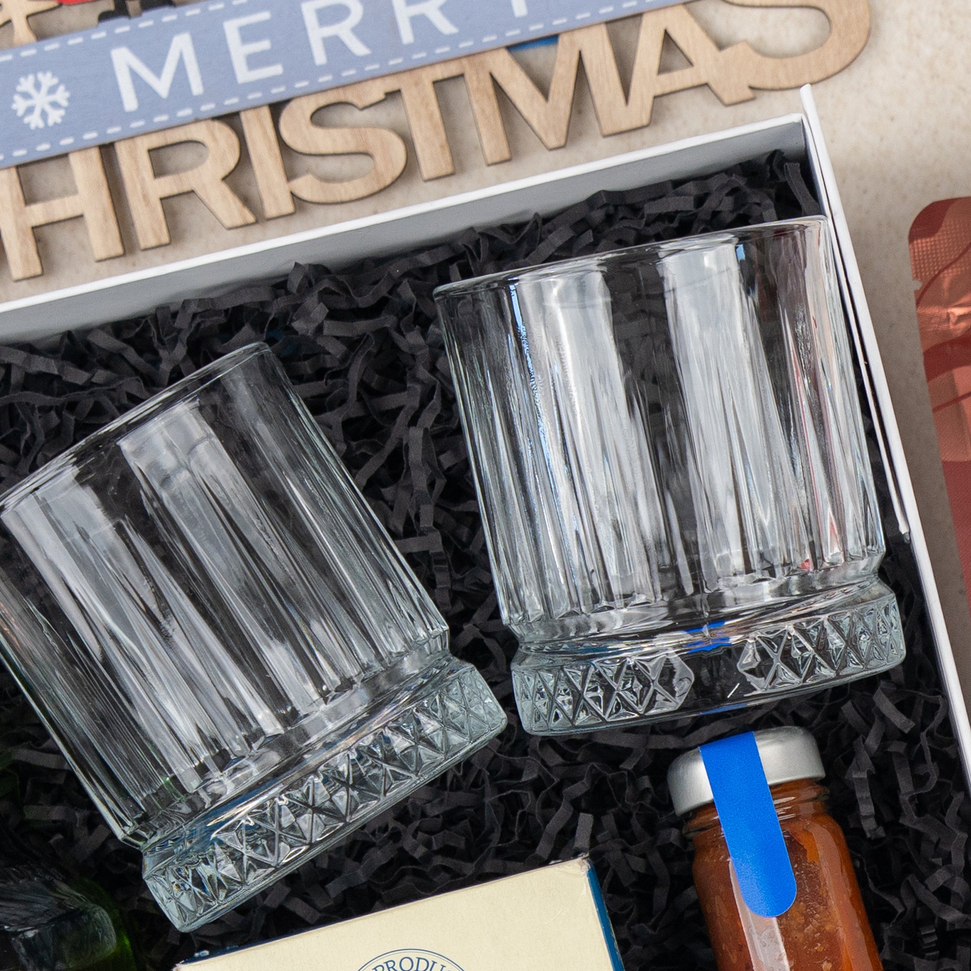 The Hamper Boutique Co's Whisky Wonder gift box includes two clear faceted glass tumblers, a small jar with orange liquid and blue label, black shredded paper, and a wooden “MERRY CHRISTMAS” sign.