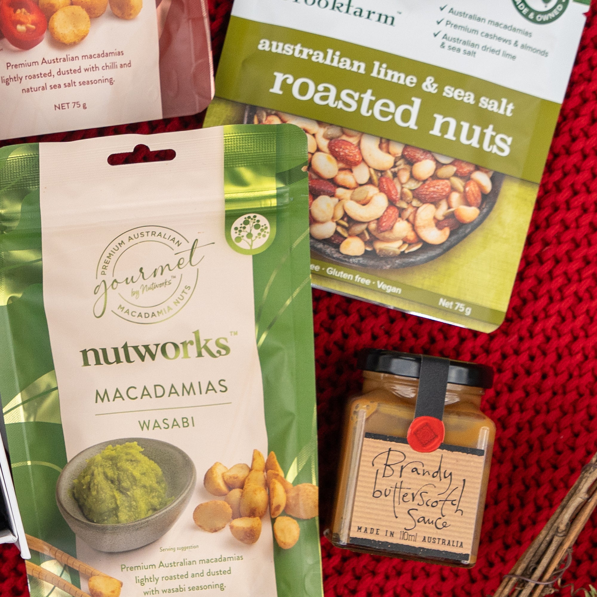 Four items from The Hamper Boutique Co are displayed: wasabi macadamias, roasted nuts with lime and sea salt, macadamia nuts, and a jar labeled Whisky Wonder, all arranged on a red woven surface.
