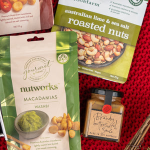 Four items from The Hamper Boutique Co are displayed: wasabi macadamias, roasted nuts with lime and sea salt, macadamia nuts, and a jar labeled Whisky Wonder, all arranged on a red woven surface.