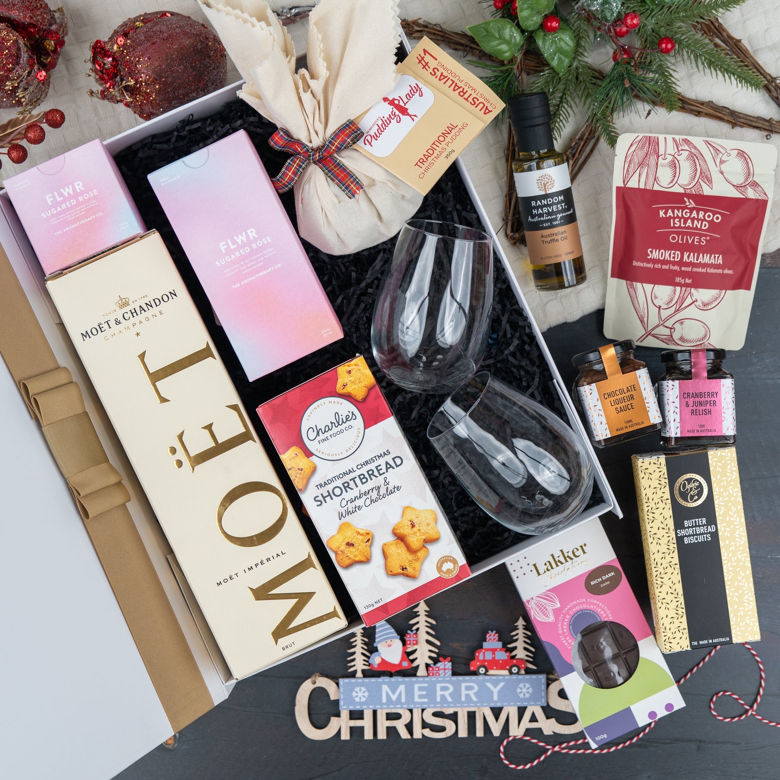 The Hamper Boutique Co's Luxurious Celebrations features Moët & Chandon, chocolate, shortbread, smoked olives, tea, wine glasses, and festive décor in a gift box arranged with a Merry Christmas sign.
