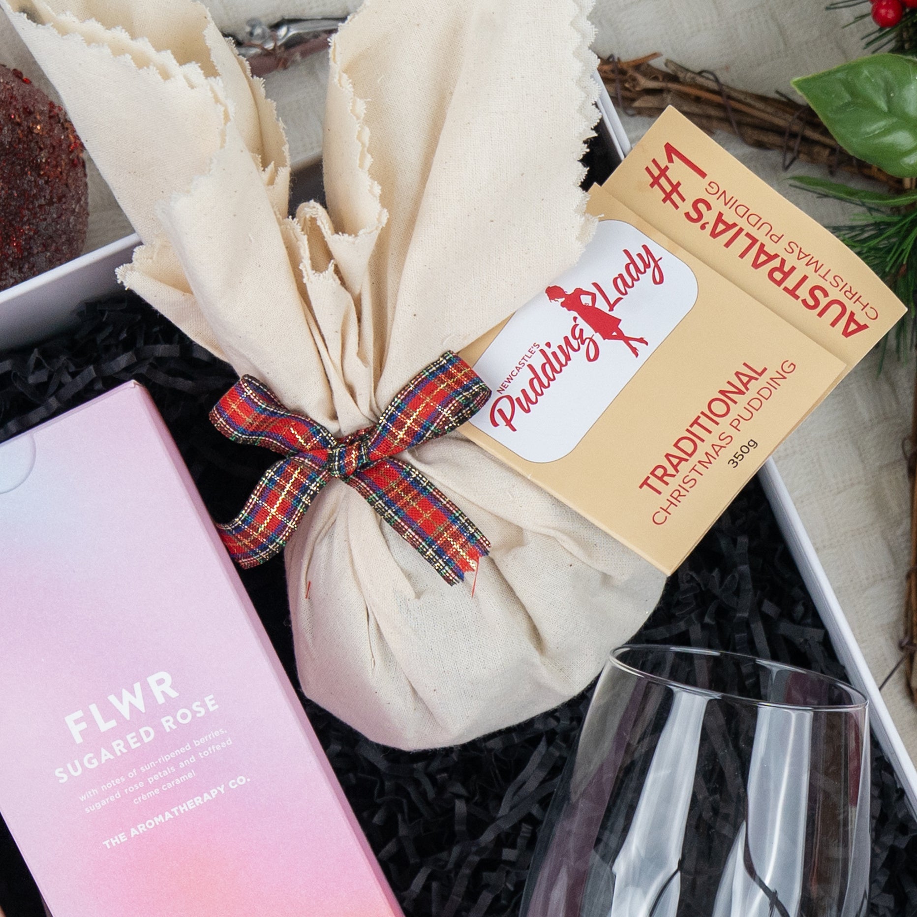 The Hamper Boutique Co’s Luxurious Celebrations gift box features a fabric-wrapped Christmas pudding with plaid ribbon, a FLWR Sugared Rose pink box, a wine glass, and festive sprigs of berries and greenery.