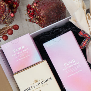 The Hamper Boutique Co's Luxurious Celebrations includes two FLWR Sugared Rose aromatherapy products, Moët & Chandon champagne, red sequin ornaments, and a cream plaid-ribbon cloth.