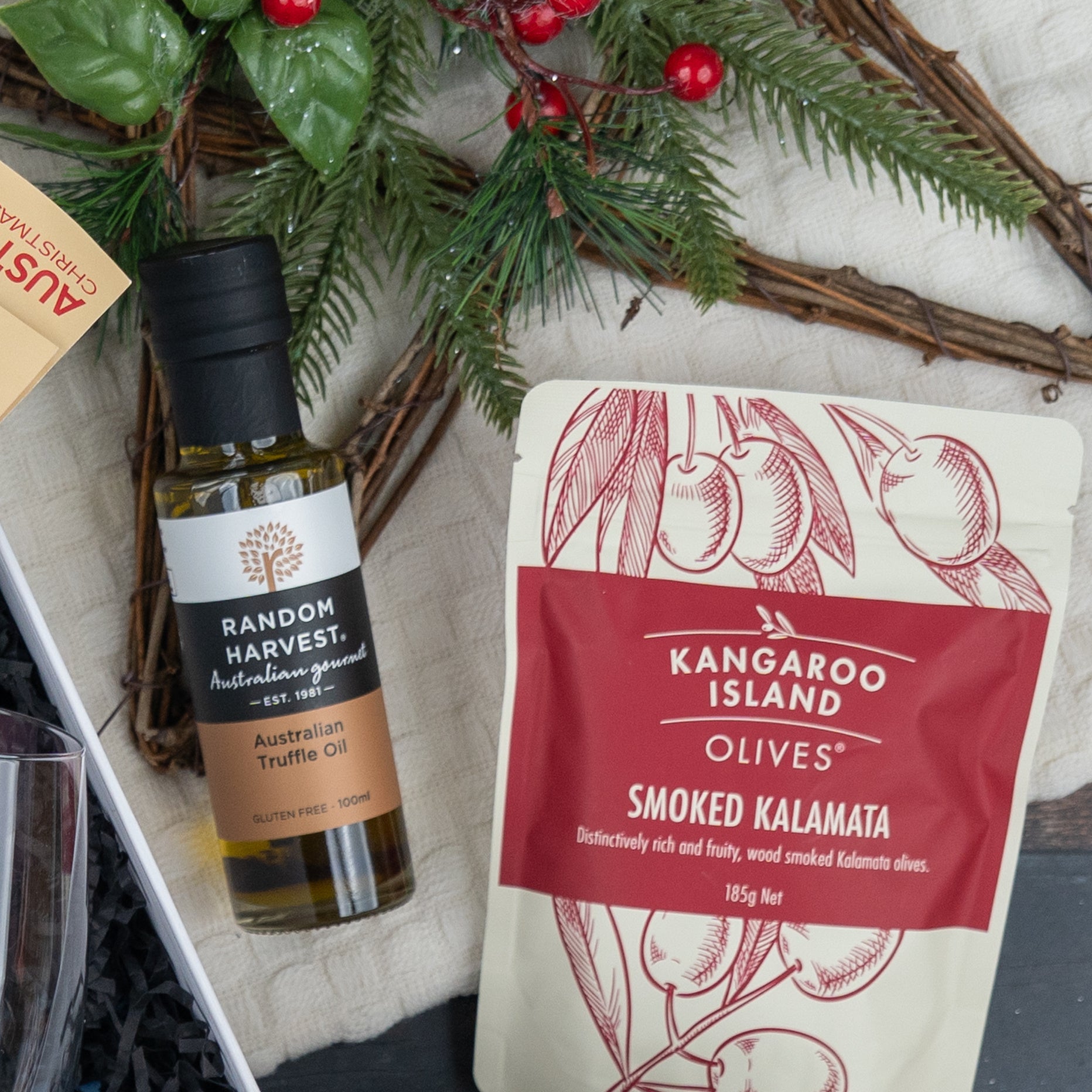 The Hamper Boutique Co's Luxurious Celebrations hamper features truffle oil and smoked Kalamata olives, elegantly arranged on a white cloth with festive greenery and red berries.