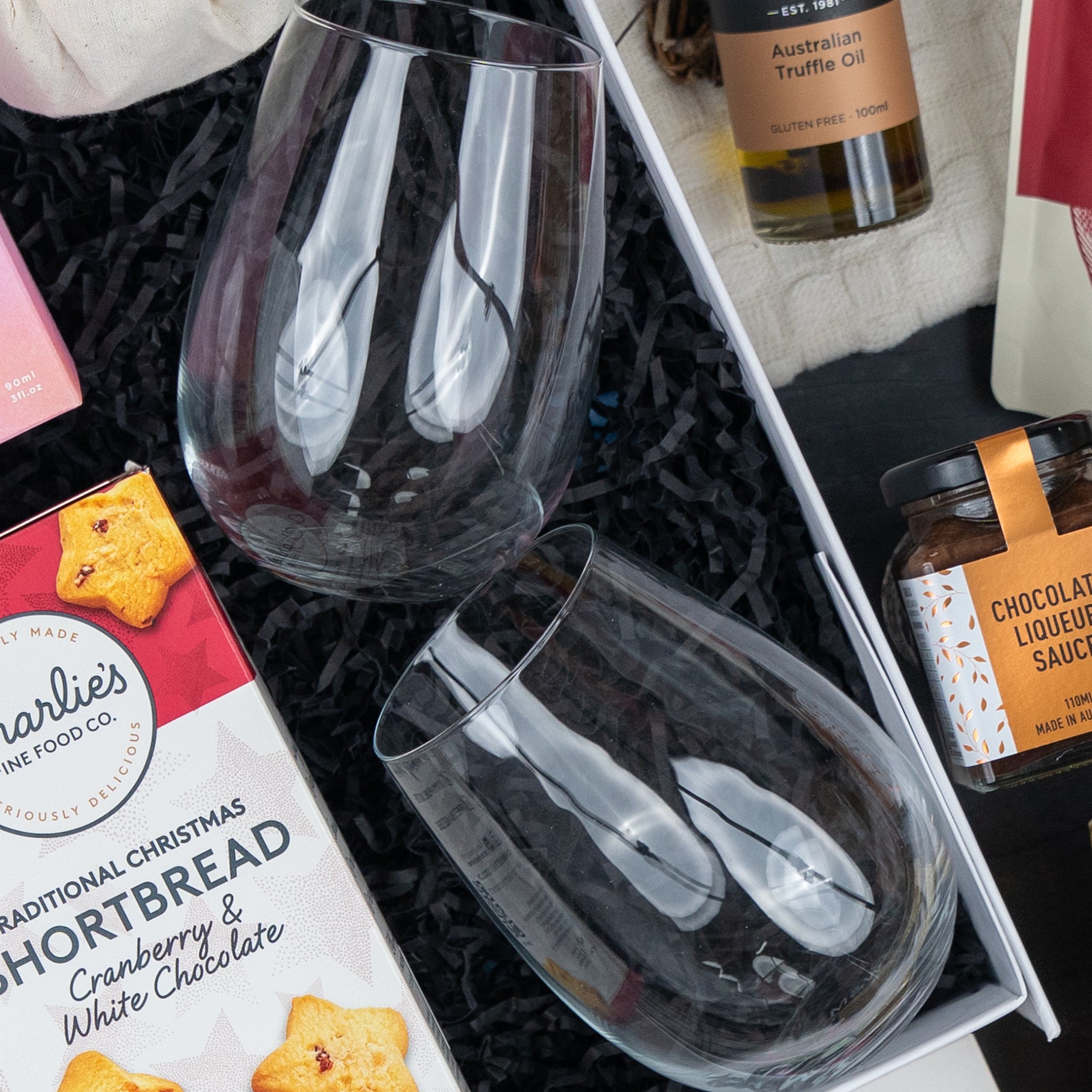 The Hamper Boutique Co's Luxurious Celebrations gift box features two empty stemless wine glasses, cranberry shortbread cookies, chocolate sauce, and truffle oil nestled in black shredded paper.