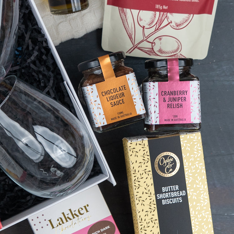 The Hamper Boutique Co’s Luxurious Celebrations gift box features two wine glasses, chocolate liqueur sauce, cranberry & juniper relish, and butter shortbread biscuits arranged elegantly on a dark surface.