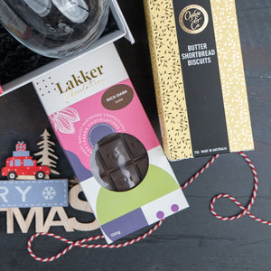 A Luxurious Celebrations gift box by The Hamper Boutique Co is displayed on a dark surface, with part of a Merry Christmas sign and a red-and-white striped string nearby.
