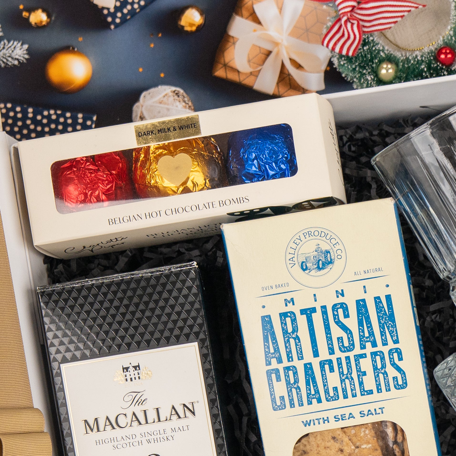 The Hamper Boutique Co's Whisky Celebration Extravaganza features Belgian hot chocolate bombs, mini artisan crackers, The Macallan scotch whisky, a glass tumbler, and festive decor on black shredded paper—perfect for gifting.