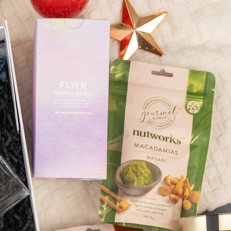 A box labeled "Whisky Celebration Extravaganza" by The Hamper Boutique Co rests beside a gold star ornament and a red object on a textured cream surface.