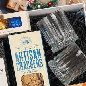 The Hamper Boutique Co's Whisky Celebration Extravaganza features two empty glass tumblers, mini artisan crackers, chocolate truffles, and festive holiday decorations arranged on shredded black paper.