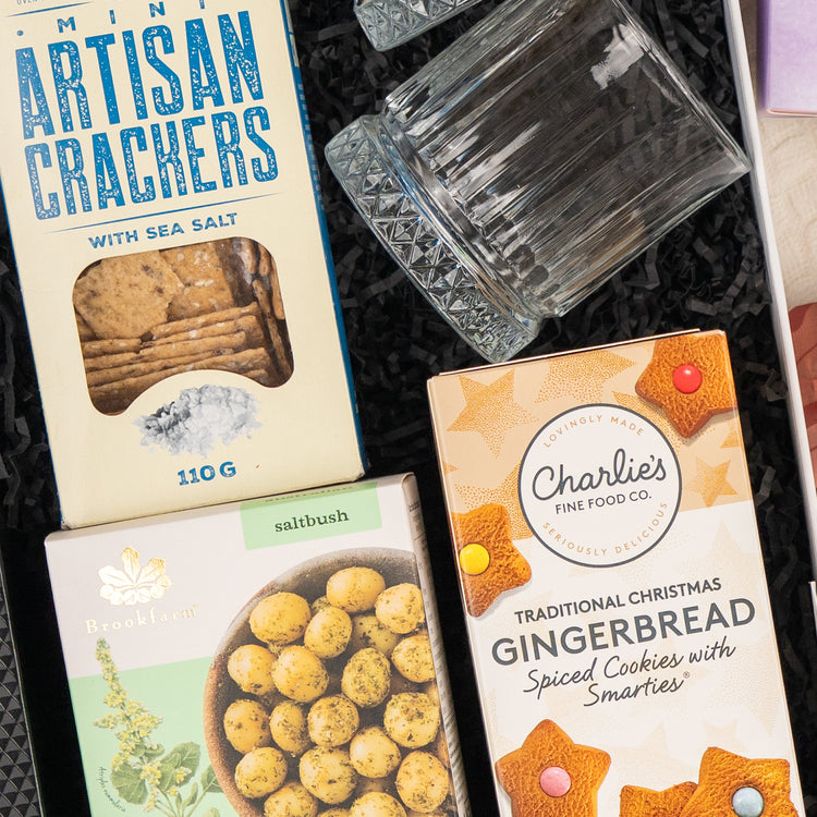 A close-up of The Hamper Boutique Co's Whisky Celebration Extravaganza, featuring mini artisan crackers, a glass tumbler, Brookfarm saltbush macadamias, and Charlie’s gingerbread cookies arranged on black shredded paper.