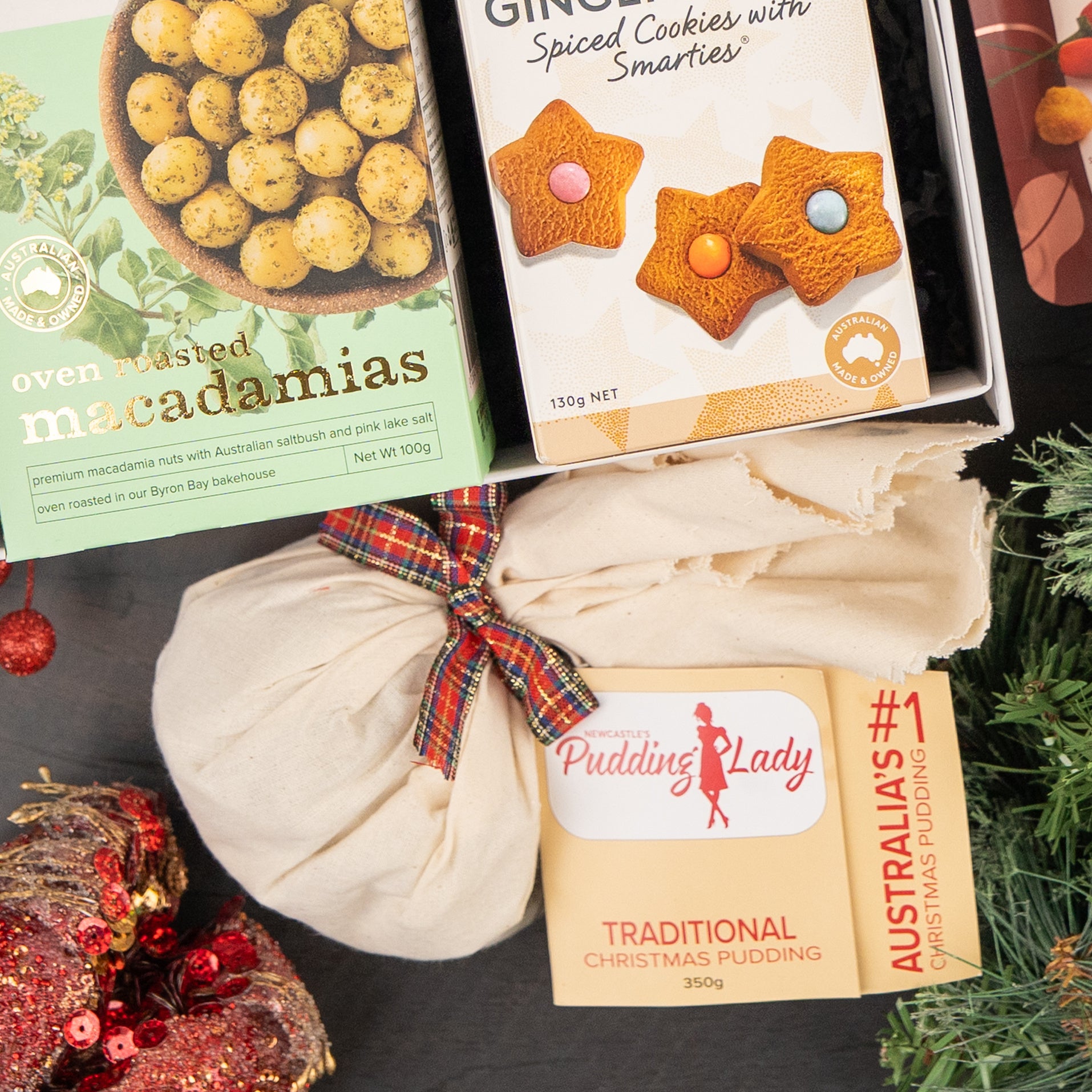 A festive flat lay featuring The Hamper Boutique Co Whisky Celebration Extravaganza, showcasing oven roasted macadamias, ginger spiced Smarties cookies, and a tartan-wrapped traditional Christmas pudding amidst holiday greenery.