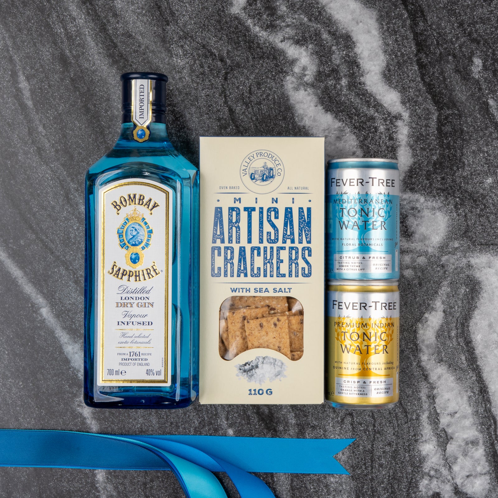 The Hamper Boutique Co’s Bombay Sapphire Gin and Tonic Hamper is displayed on a gray marble surface with a blue ribbon in the corner.