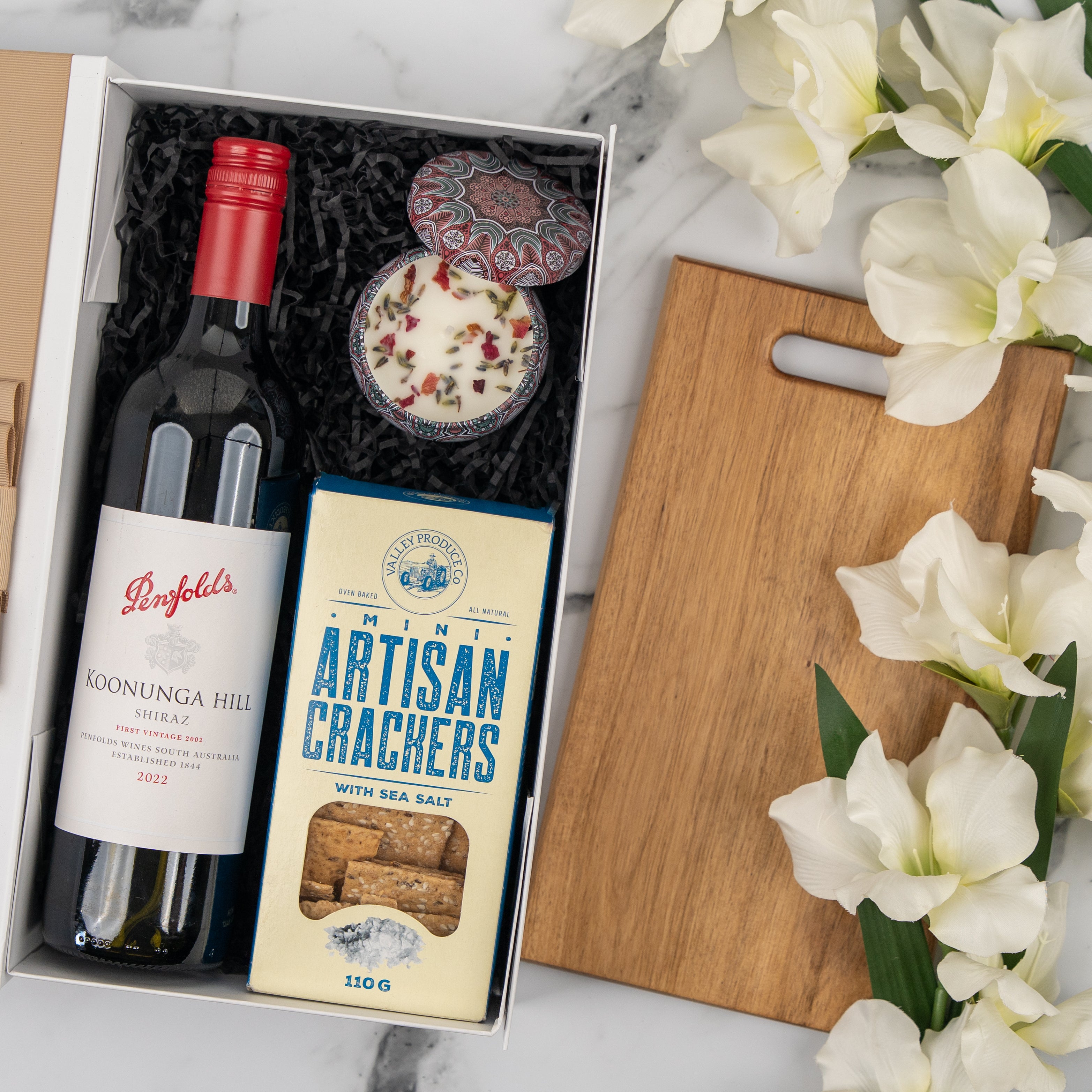 The Hamper Boutique Co’s Wine, Candle and Board Hamper features a bottle of wine, artisan crackers, a decorative candle, a wooden serving board, and white flowers—making it the perfect elegant gift set.
