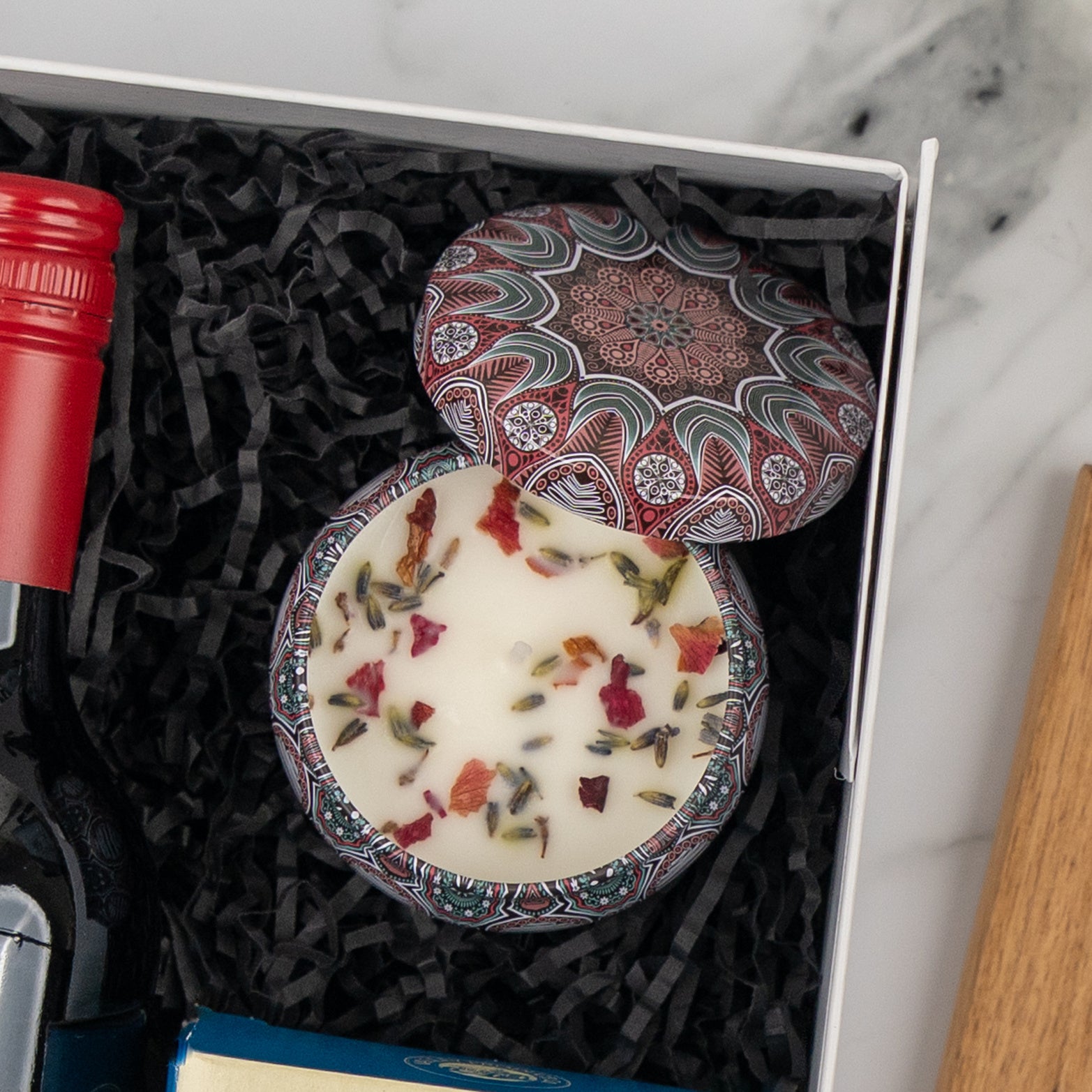 The Hamper Boutique Co's Wine, Candle and Board Hamper includes a decorative flower-topped tin candle in a gift box with black paper shreds, alongside a red wine bottle and another item, arranged on a marble surface.