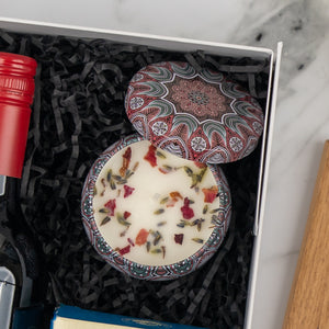 The Hamper Boutique Co's Wine, Candle and Board Hamper includes a decorative flower-topped tin candle in a gift box with black paper shreds, alongside a red wine bottle and another item, arranged on a marble surface.