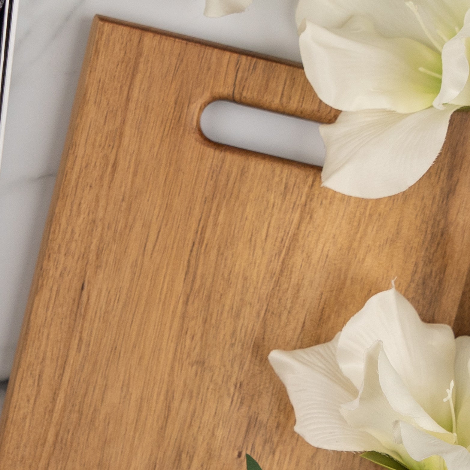 The Hamper Boutique Co’s Wine, Candle and Board Hamper features a light wooden cutting board with a handle slot, elegantly displayed on a white surface next to two large white flowers.