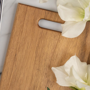 The Hamper Boutique Co’s Wine, Candle and Board Hamper features a light wooden cutting board with a handle slot, elegantly displayed on a white surface next to two large white flowers.