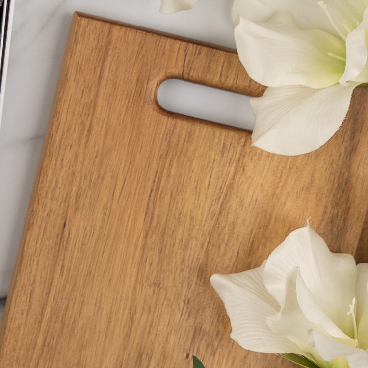 The Hamper Boutique Co’s Wine, Candle and Board Hamper features a light wooden cutting board with a handle slot, elegantly displayed on a white surface next to two large white flowers.