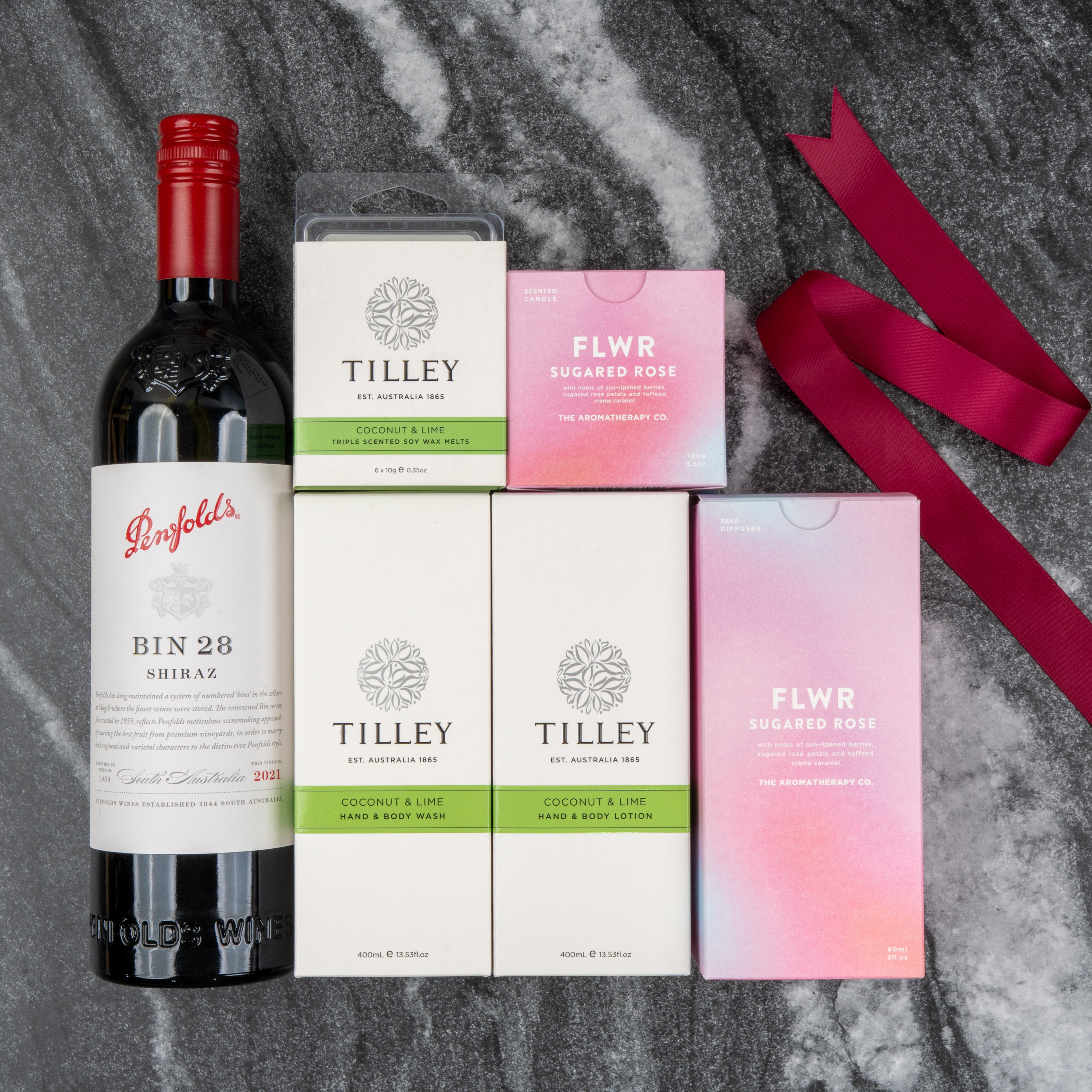 The Hamper Boutique Co's Penfolds Spa and Indulgence Hamper features Penfolds Bin 28 Shiraz wine, Tilley Coconut & Lime soap, hand & body lotion, a candle, and two FLWR Sugared Rose candles arranged on gray marble with a pink ribbon.